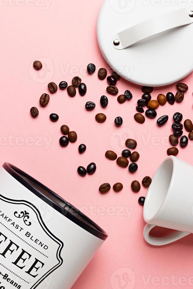 Coffee beans out of the coffee pot with the pot lid next to it and a white cup on a pink background. photo