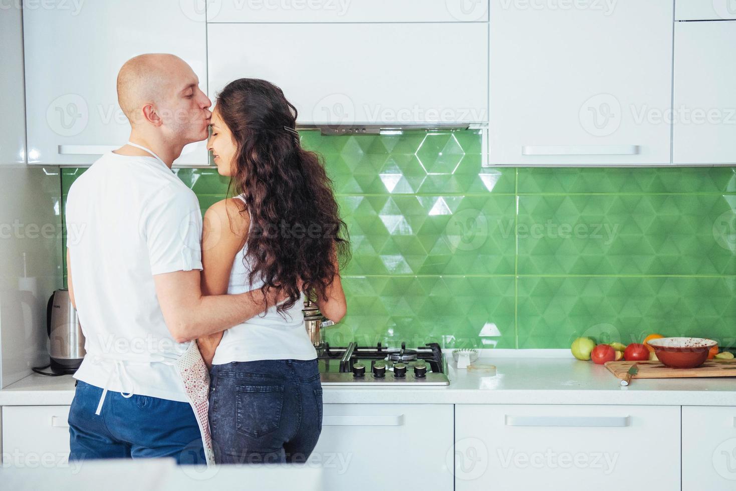 Beautiful young couple is talking, looking at camera and smiling while cooking in kitchen. photo