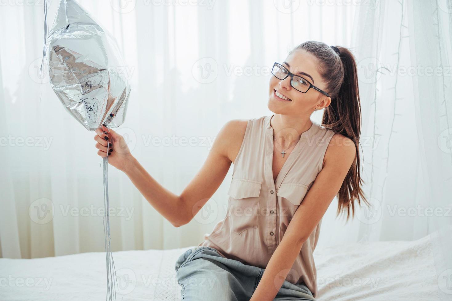 Closeup cute brunette girl in the studio, smiling broadly and playing with transparent and silver balloons. She wears glasses and twisted hair. photo