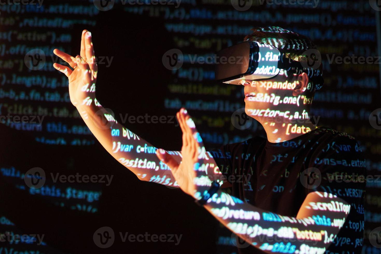 la doble exposición de un hombre caucásico y un casco de realidad virtual es presumiblemente un jugador o un pirata informático que descifra el código en una red o servidor seguro, con líneas de código foto