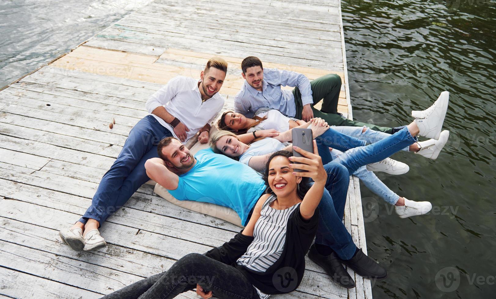 un grupo de jóvenes sentados en el borde del muelle y haciendo selfie. amigos disfrutando de un juego en el lago foto