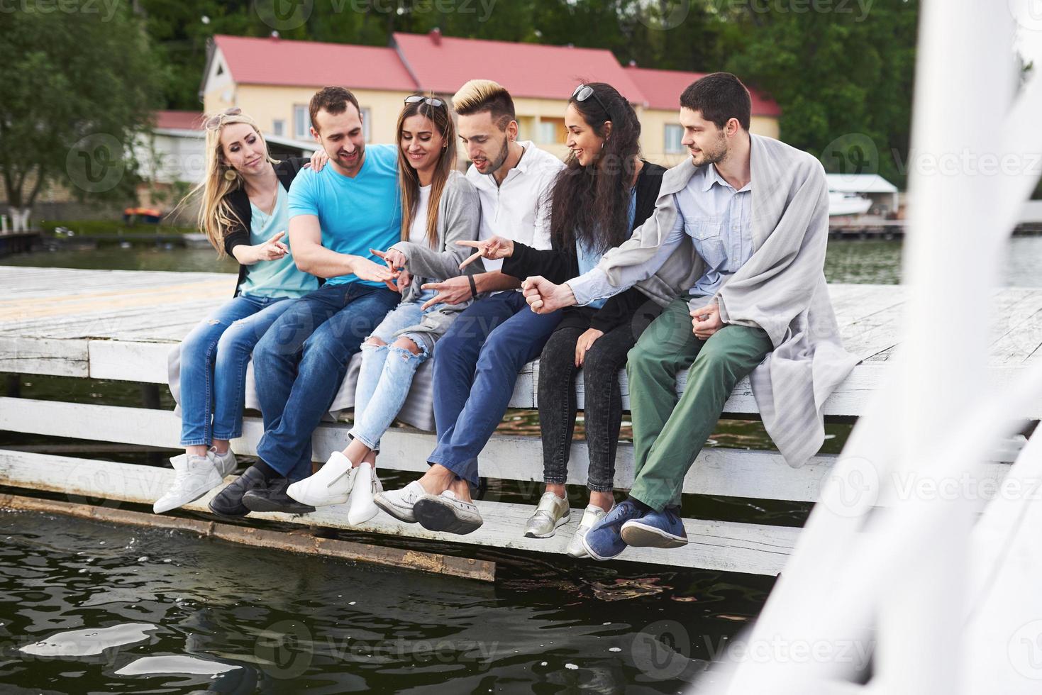 Portrait of joyful group of young people sitting on the edge of the pier, outdoors in nature. Friends enjoying a day on the lake. photo