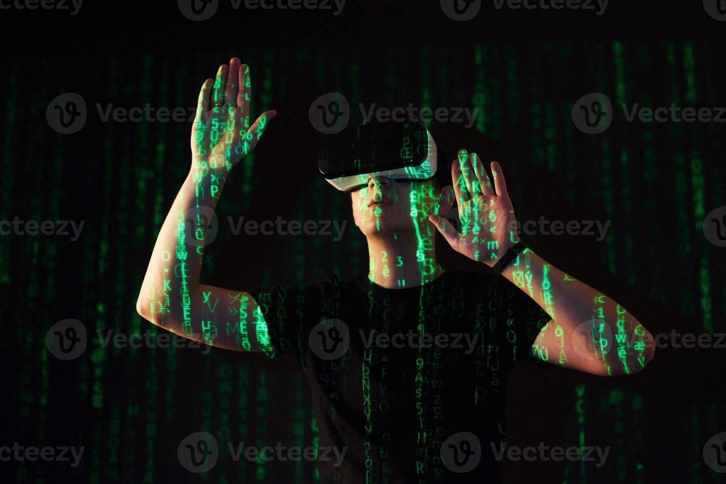 la doble exposición de un hombre caucásico y un casco de realidad virtual es presumiblemente un jugador o un pirata informático que descifra el código en una red o servidor seguro, con líneas de código foto