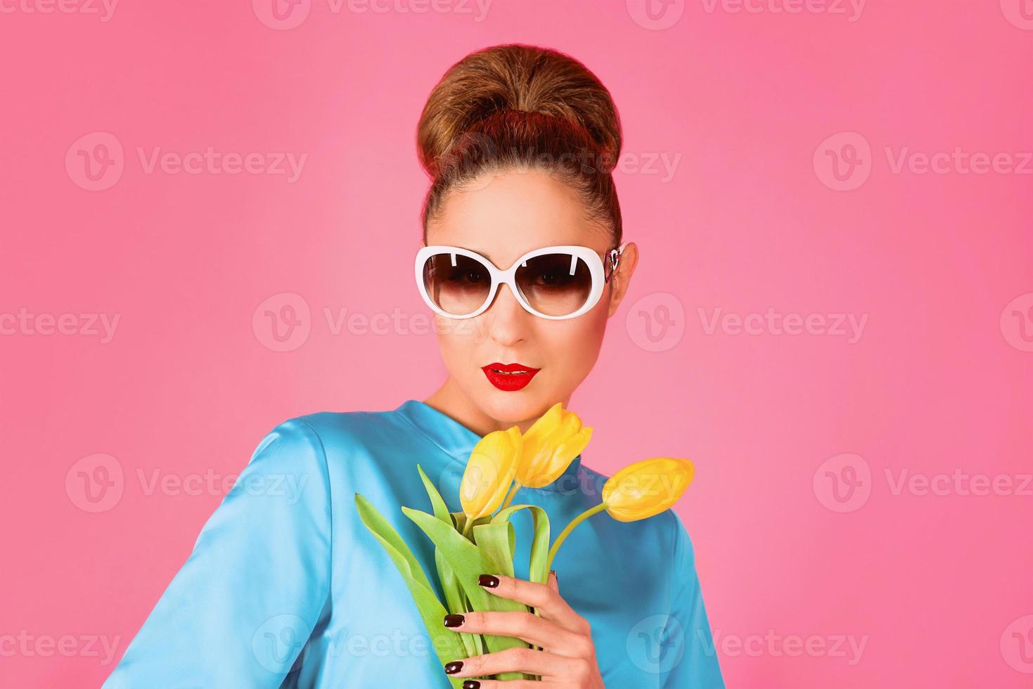 retrato de una joven hermosa mujer vestida de seda azul y gafas de sol blancas con tulipanes amarillos en el fondo rosa foto