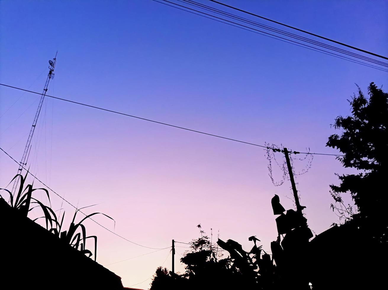 foto de silueta de casas, postes de electricidad y plantas al amanecer en el cielo azul, violeta y rosa del pueblo.