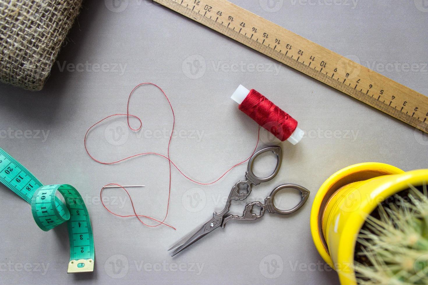 Sewing accessories scissors, measuring tape, ruler, fabric. In center of composition - red thread, laid out in the shape of a heart, with a needle at the end. With cactus for home furnishings photo