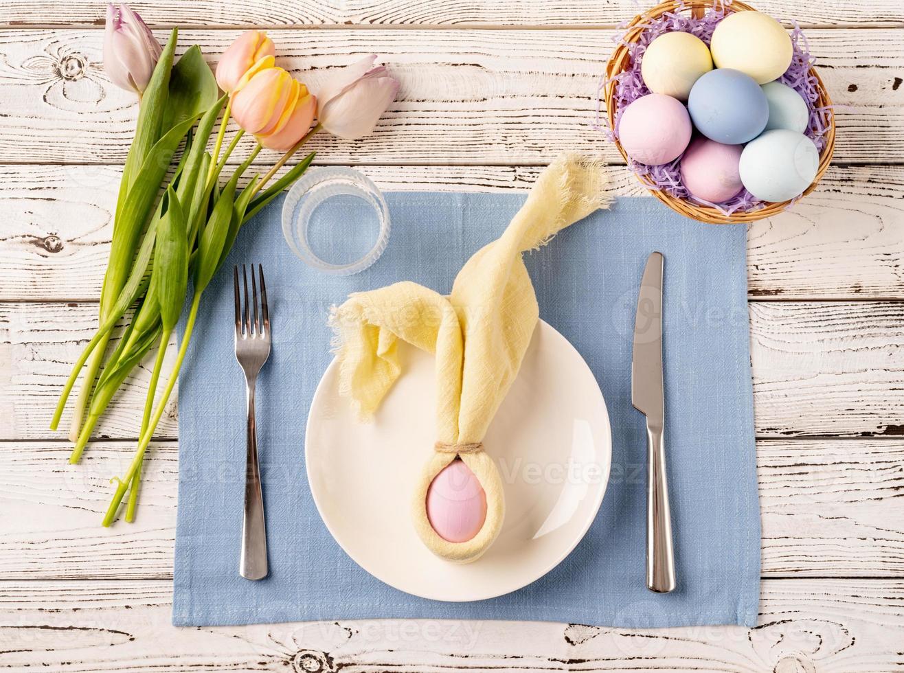 Table setting for Easter feast. Easter eggs, bunny, tulips and cutlery top view flat lay on white rustic background photo