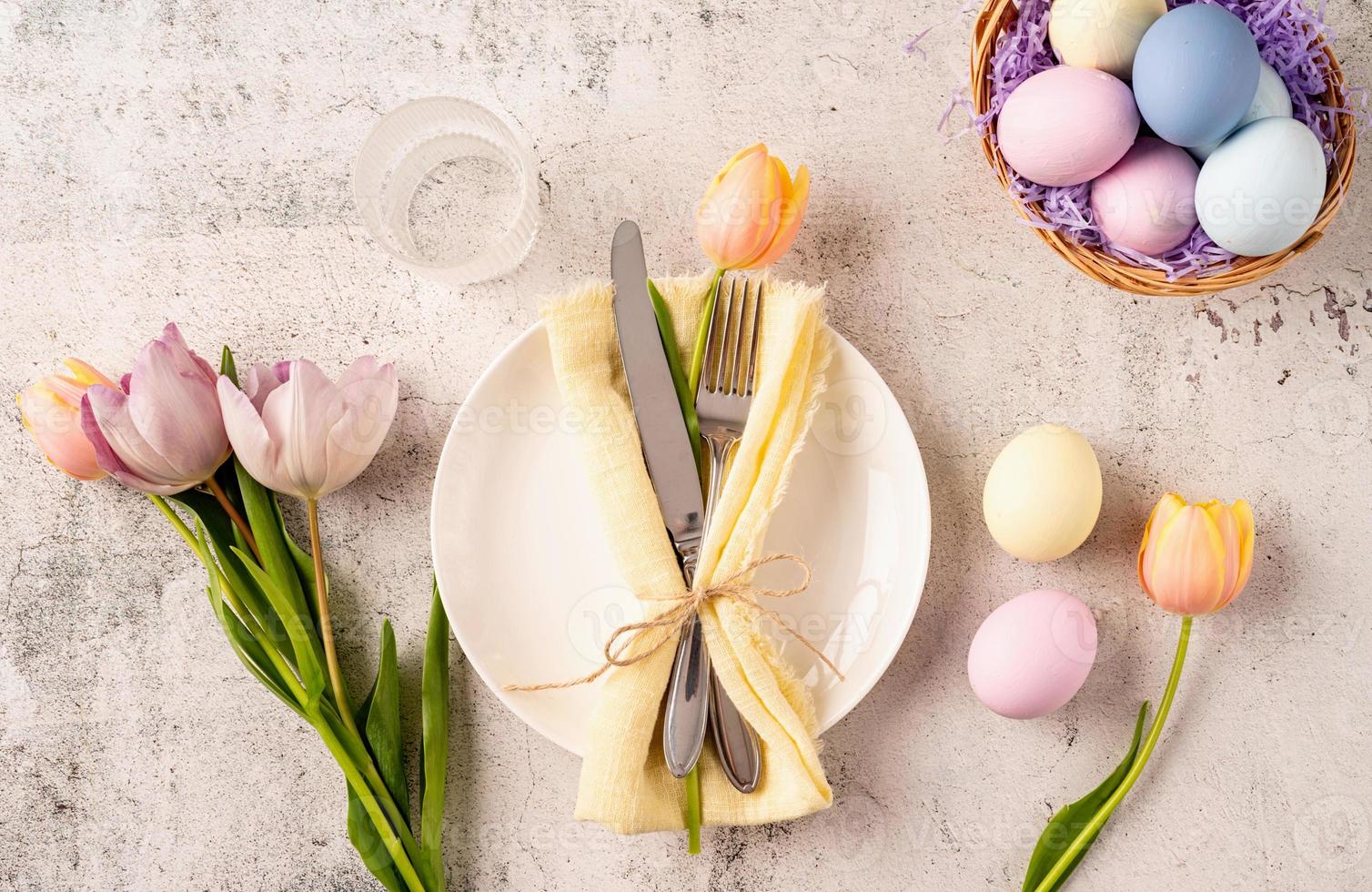 Table setting for Easter feast. Easter eggs, bunny, tulips and cutlery top view flat lay on concrete background photo