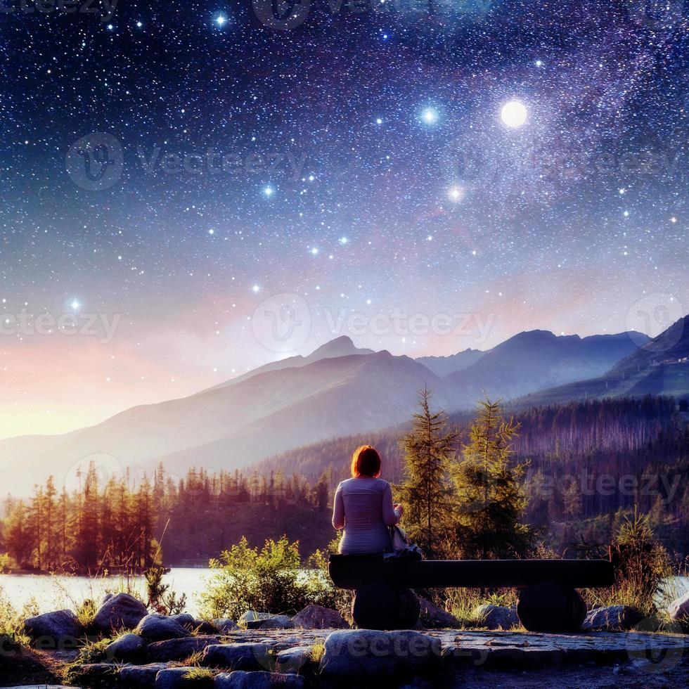 Lake Strbske pleso in High Tatras mountain, Slovakia, Europe. Fantastic starry sky and the milky way. The girl sits on a bench and looks into the fairy-tale sky. photo