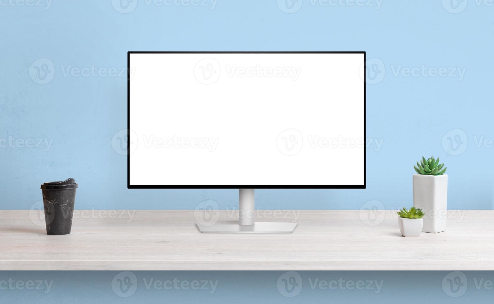 Computer display on work desk with isolated screen for web page presentation. Coffee and plants beside. Blue wall in background photo