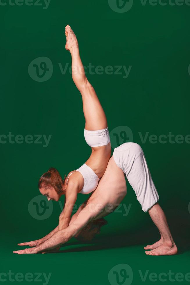 Beautiful sporty woman and man in white clothes doing yoga asanas together indoor on green background photo