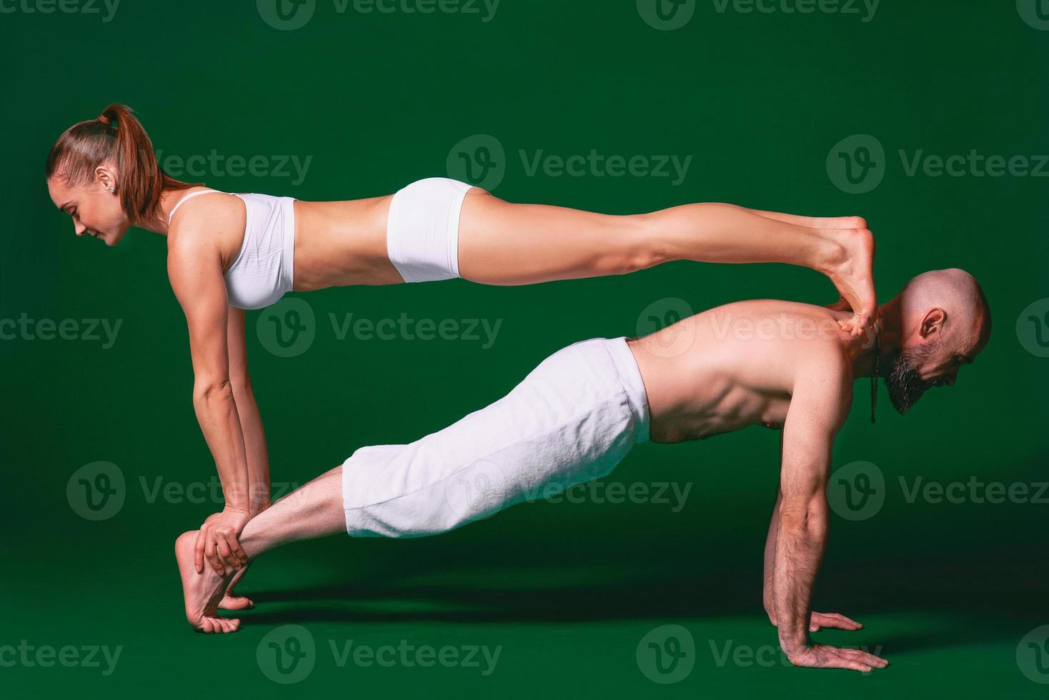 Beautiful sporty woman and man in white clothes doing yoga asanas together indoor on green background photo
