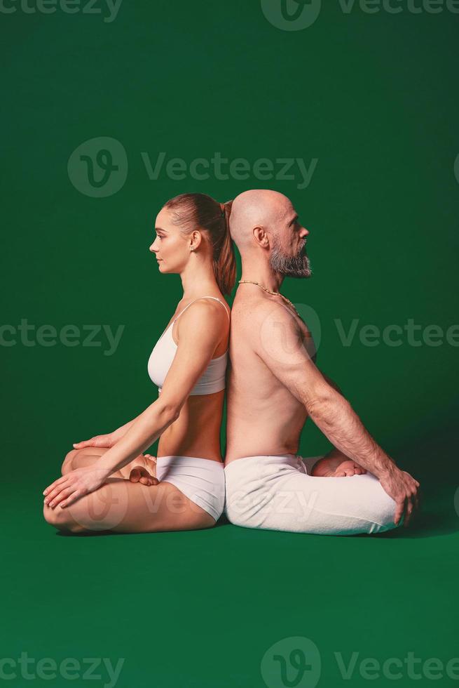 Beautiful sporty woman and man in white clothes doing yoga asanas together indoor on green background photo