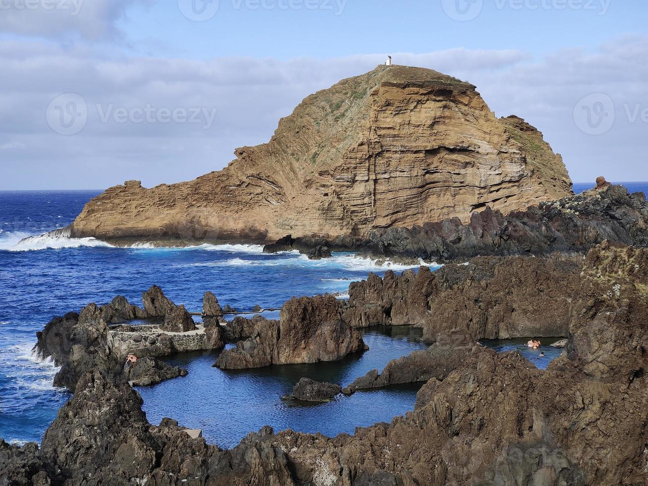 Natural pools in the middle of the Atlantic ocean in Porto Moniz, Madeira Island, Portugal. Amazing holiday times. Clouds with sun. Ocean and waves hitting the rocks. People in the water. photo