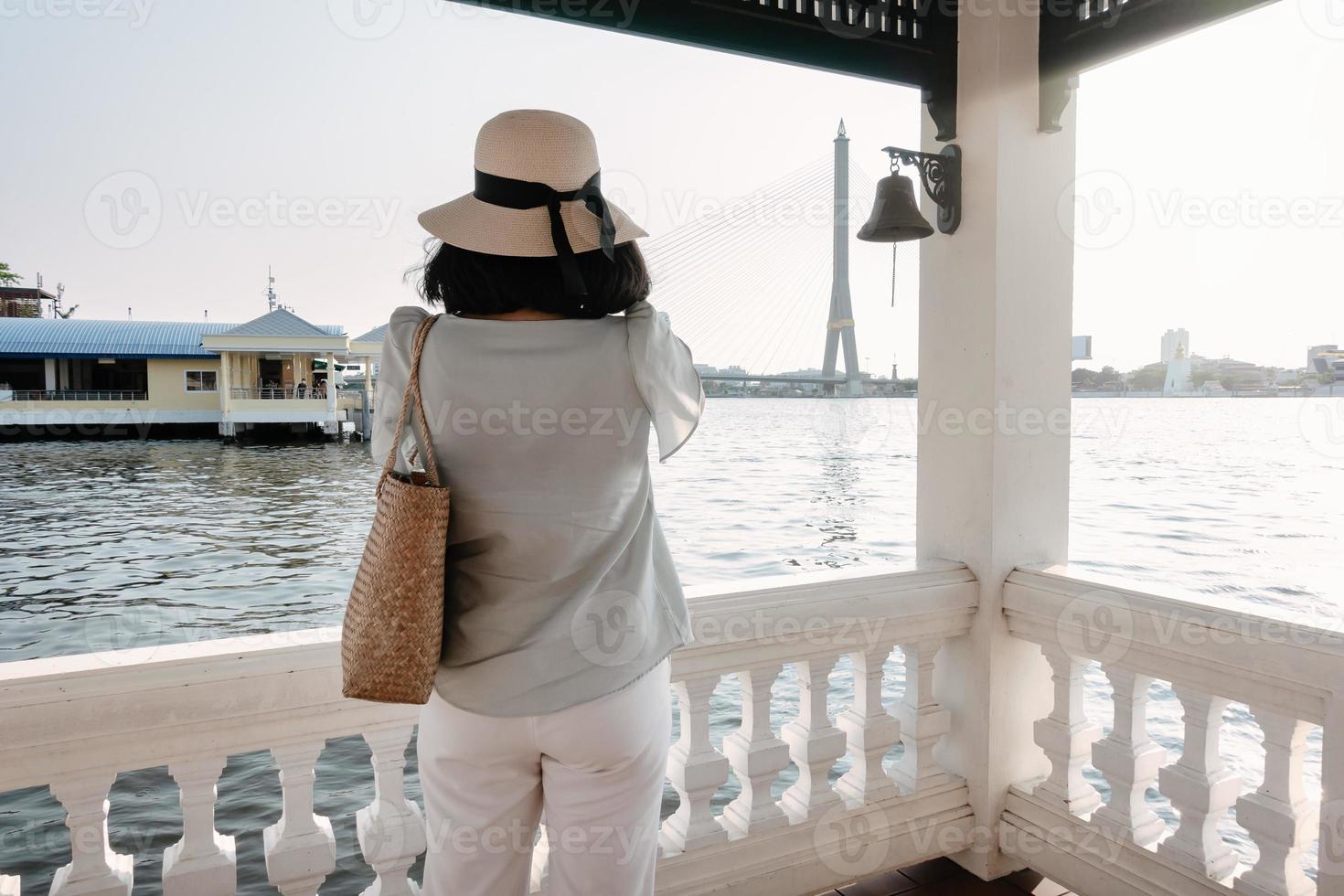 Tourist Woman Having Fun While Sightseeing Bangkok Cityscape beside The River, Rear View of Asian Tourist Woman Relaxing and Enjoy While Photographing Architecture on Her Mobile Phone. photo