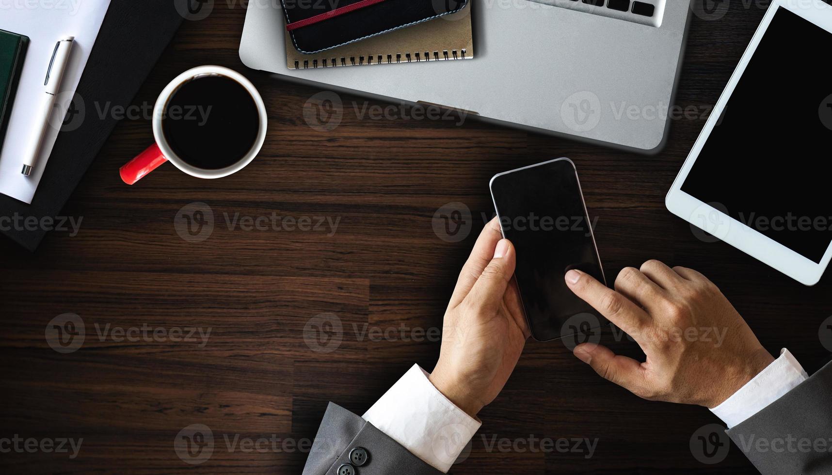 Cropped shot of male worker hands holding smartphone on dark modern office desk with office supplies photo