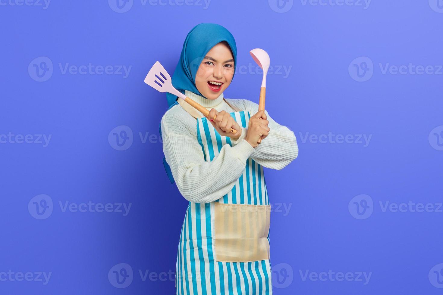 Excited beautiful Asian woman in white sweater covered in apron and hijab crossed hand hold spatula and spoon while doing housework isolated over purple background. Housekeeping concept photo