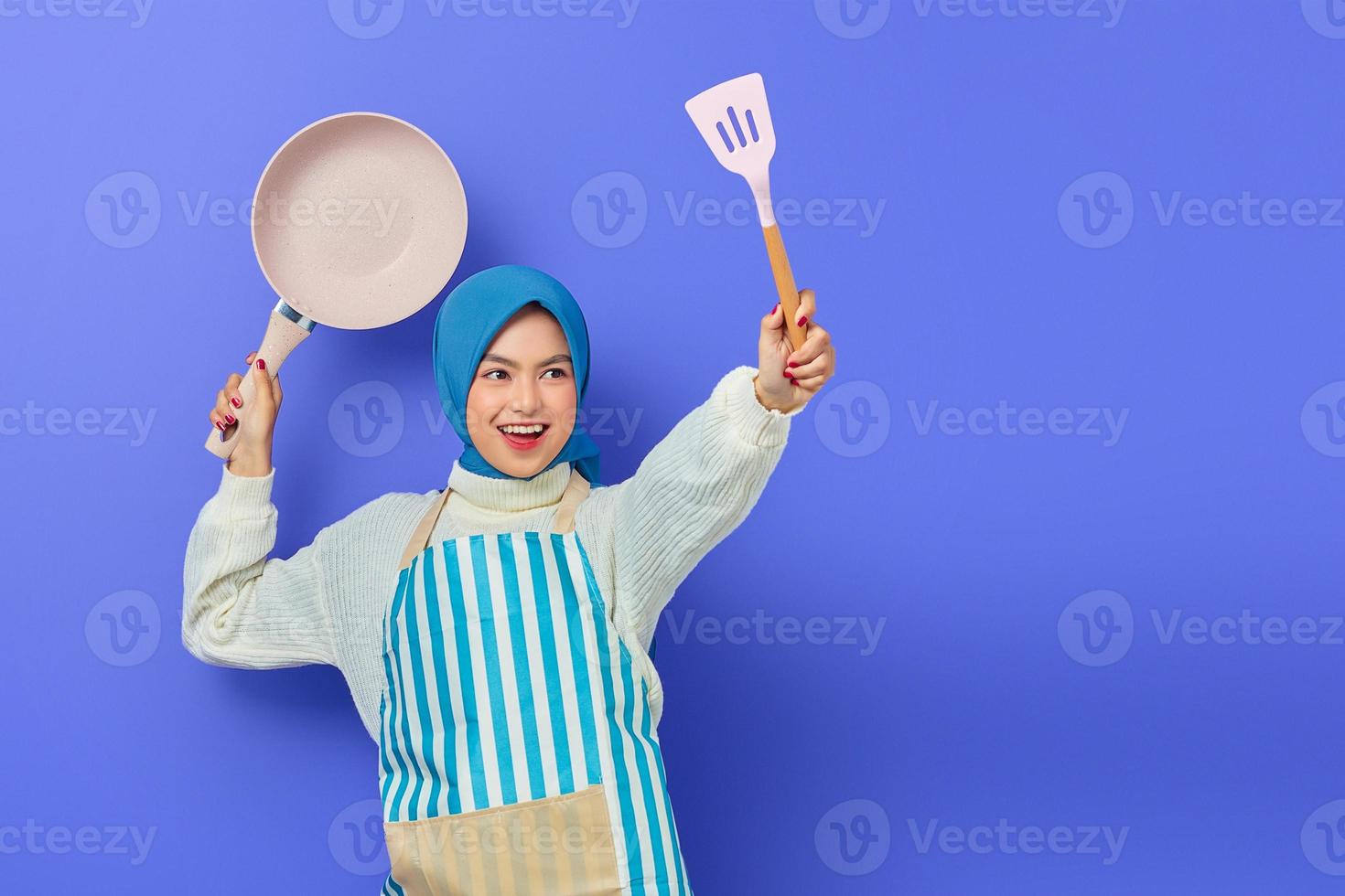 Cheerful beautiful Asian woman in white sweater covered in apron and hijab hold spatula and pan attack enemies while doing housework isolated over purple background. Housekeeping concept photo