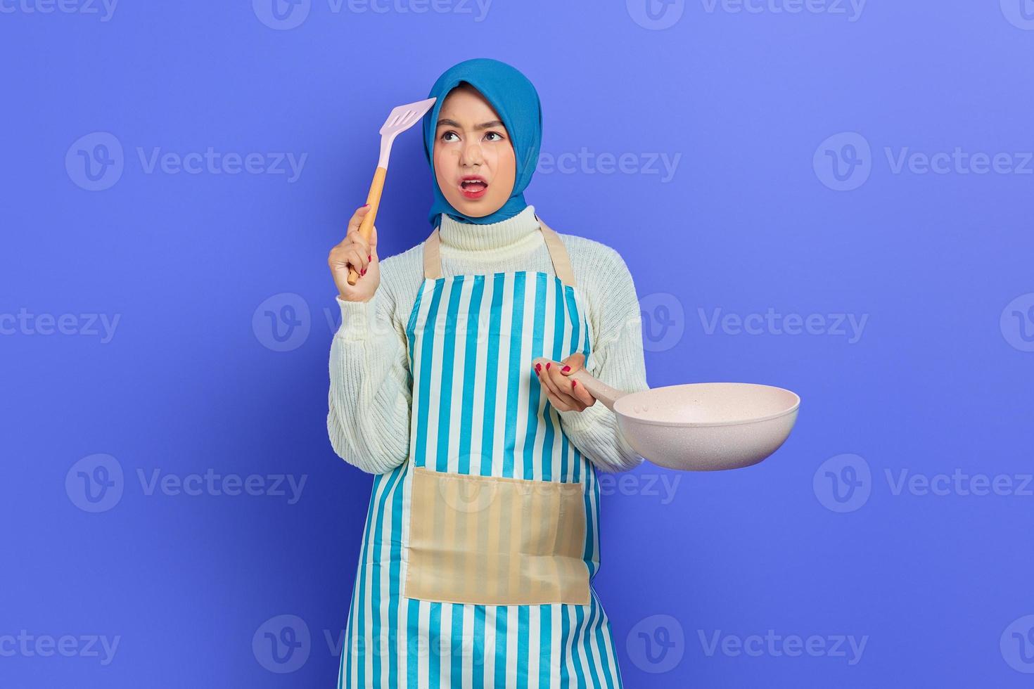 Portrait of confused young Asian woman housewife wearing apron holding frying pan and spatula, looking up thinking cooking menu isolated over purple background. Housekeeping concept photo