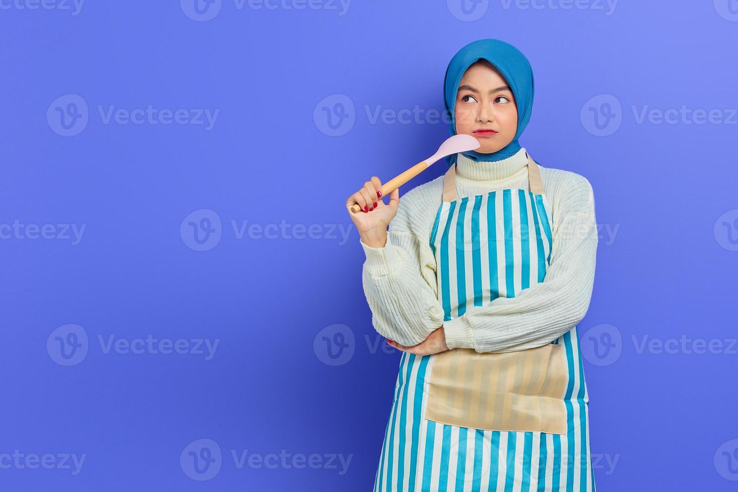 Pensive beautiful Asian woman in white sweater covered in apron and hijab holding a spoon, looking up doing housework isolated over purple background. Housekeeping concept photo