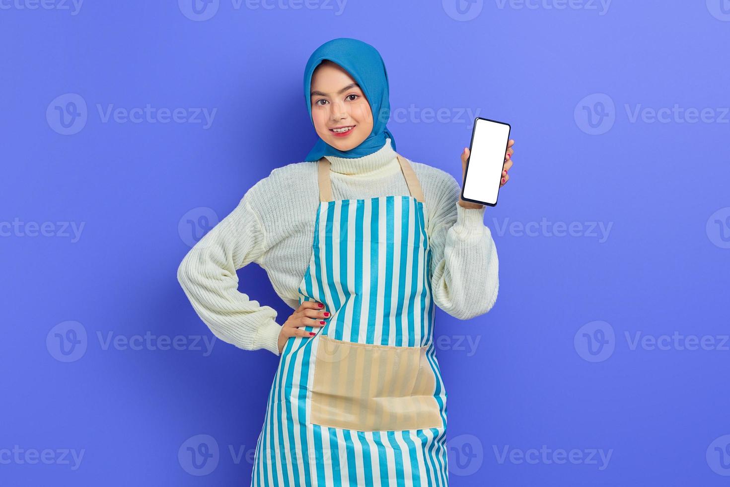 retrato de una joven musulmana asiática sonriente con hiyab y delantal sosteniendo un teléfono móvil con pantalla en blanco aislado en un fondo morado. gente ama de casa concepto de estilo de vida musulmán foto