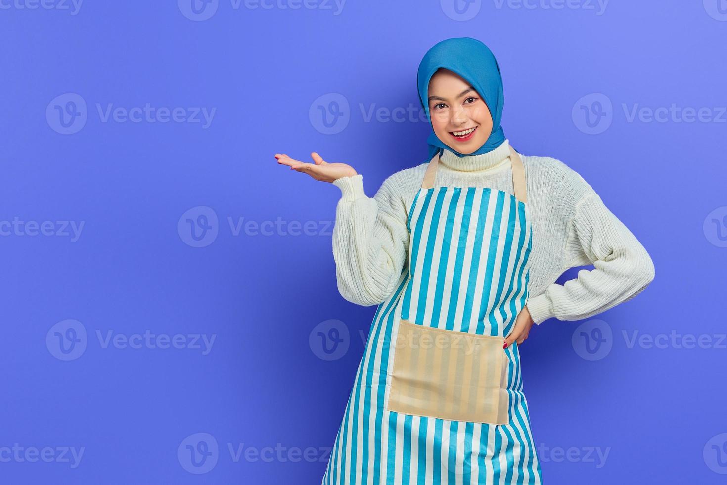 retrato de una mujer ama de casa sonriente con hiyab y delantal que muestra el producto con la mano aislada en un fondo morado. gente ama de casa concepto de estilo de vida musulmán foto