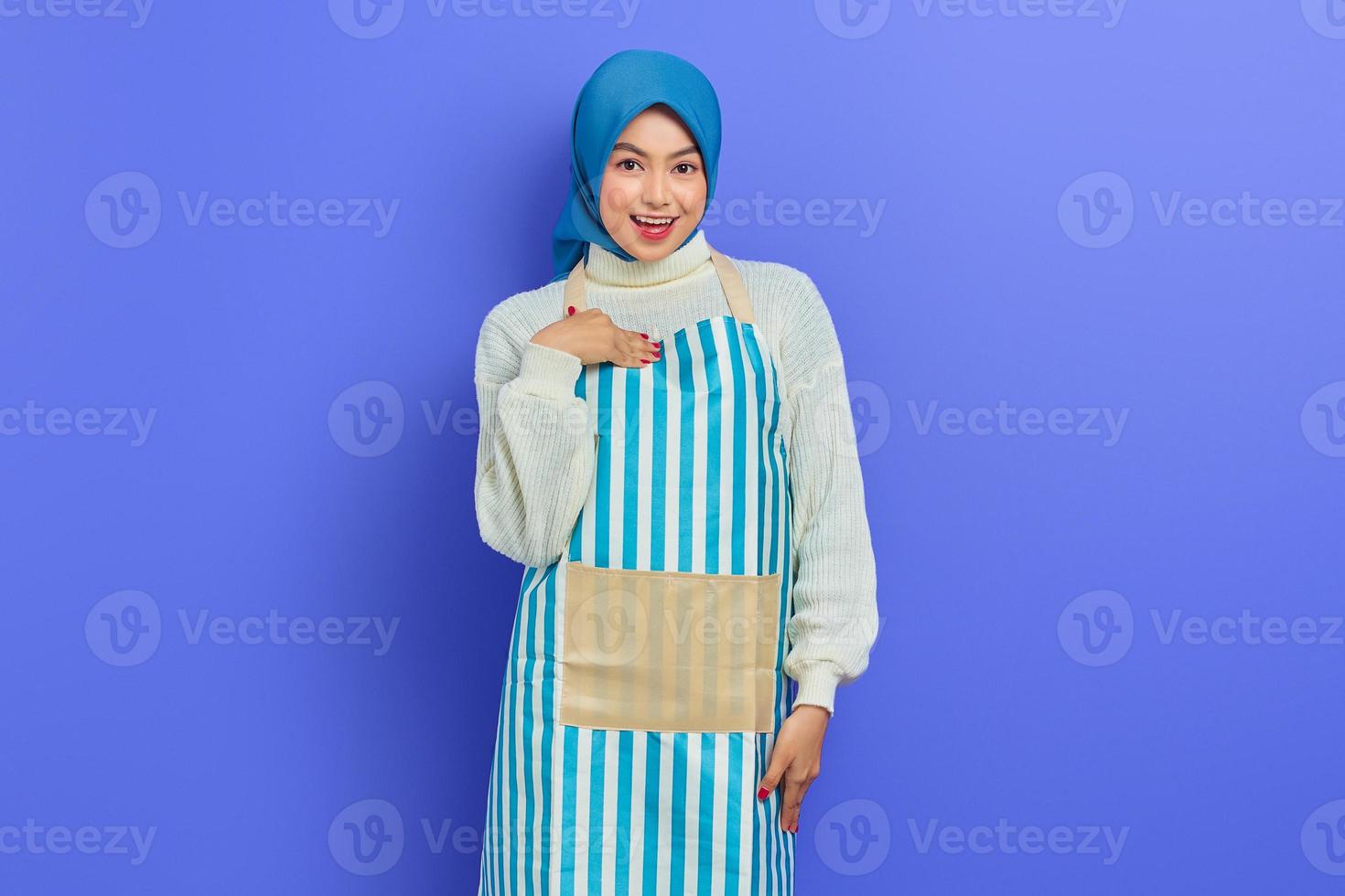 Portrait of smiling housewife woman in hijab and apron pointing at herself proudly isolated on purple background. People housewife muslim lifestyle concept photo
