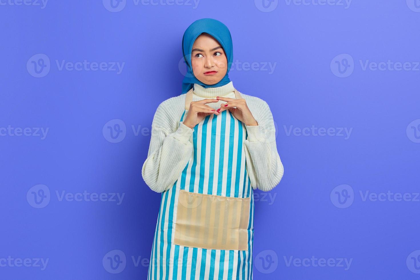 Portrait of pensive young housewife woman in hijab and apron looking seriously thinking about a question isolated on purple background. People housewife muslim lifestyle concept photo