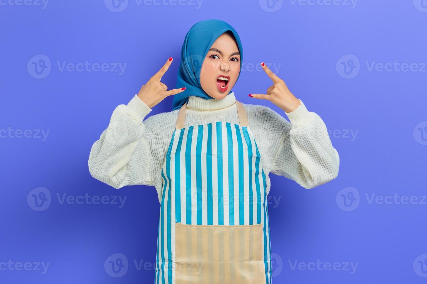 Portrait of crazy young housewife woman in hijab and striped apron, Depicting heavy metal rock sign, screaming open mouth isolated on purple background. People housewife muslim lifestyle concept photo