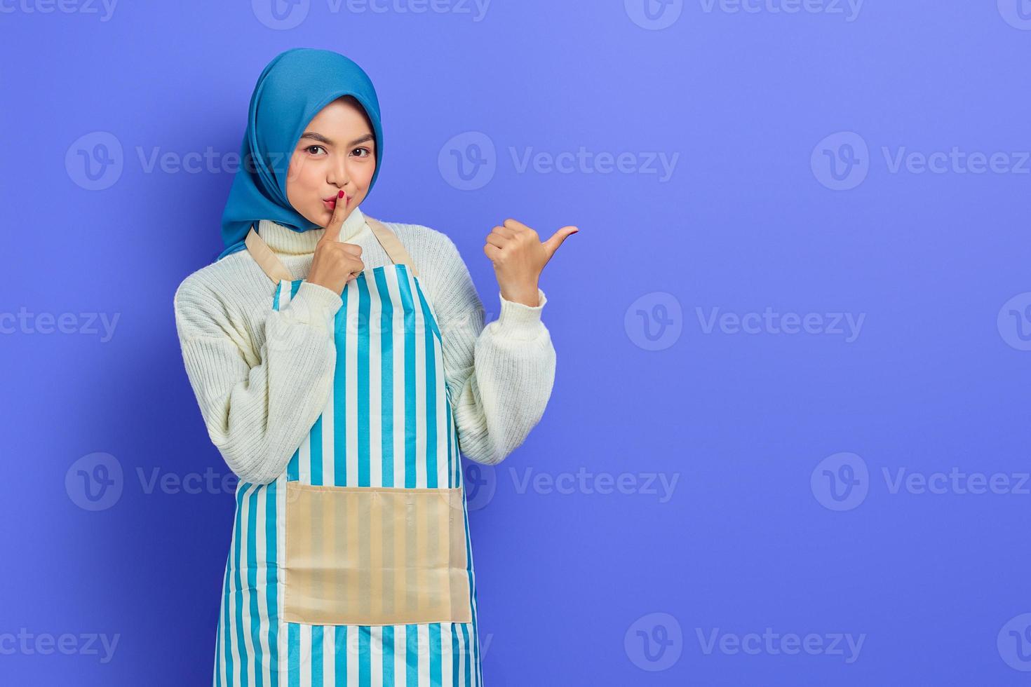 Secret young woman in hijab and stiped apron saying hush be quiet with finger on lips shhh gesture while doing housework, pointing aside with finger isolated on purple background photo