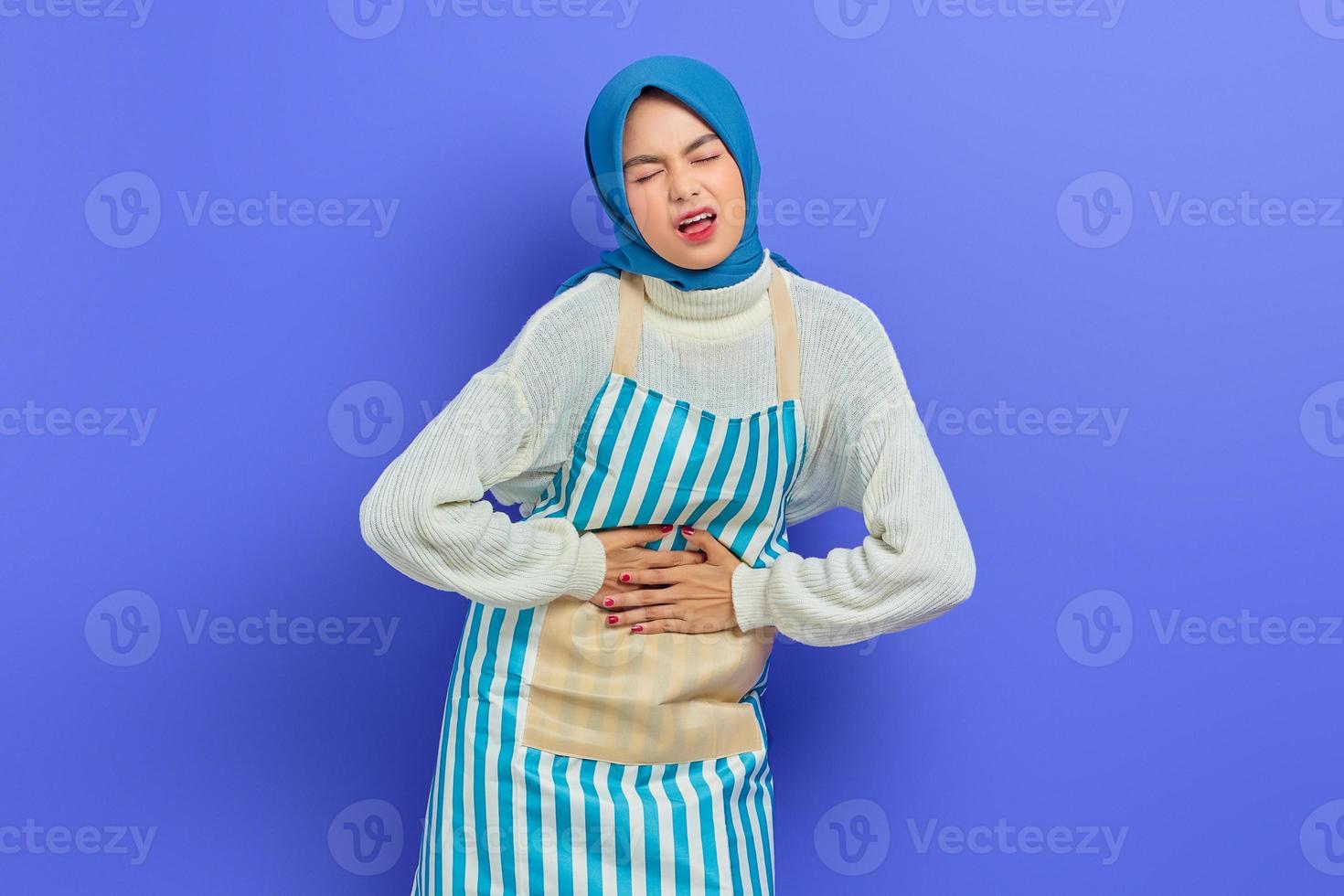 Portrait of sick young housewife woman in hijab and striped apron, Having abdominal pain, put hands on stomach on purple background. People housewife muslim lifestyle concept photo