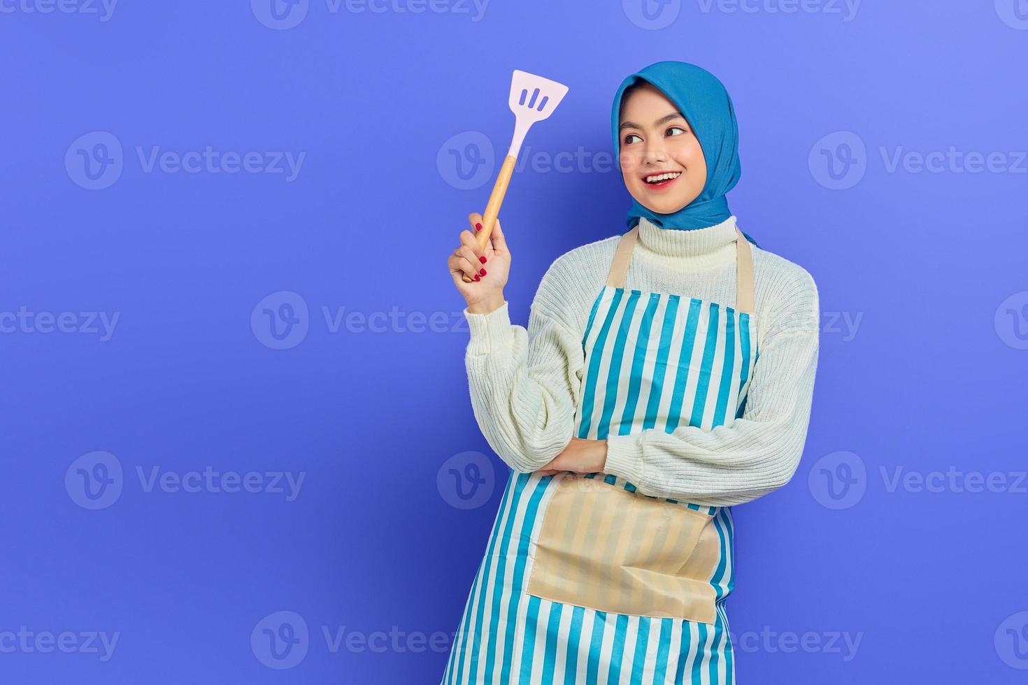 sonriente hermosa mujer asiática con suéter blanco cubierto de delantal y hiyab sosteniendo espátula, mirando hacia arriba haciendo tareas domésticas aisladas sobre fondo morado. concepto de limpieza foto