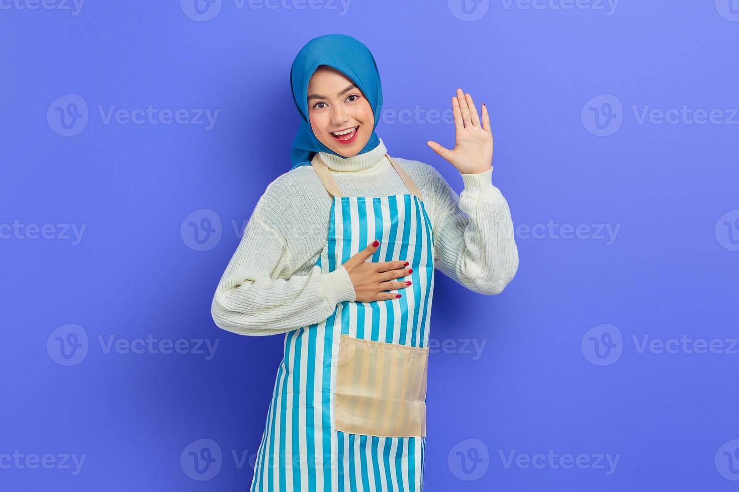 Portrait of smiling young Asian Muslim woman in hijab and striped apron, hands on chest and waving to camera isolated on purple background. People housewife muslim lifestyle concept photo