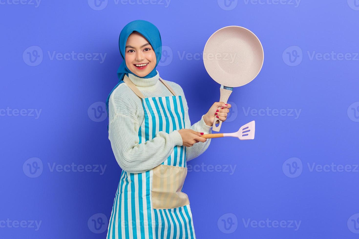 Portrait of cheerful young Asian Muslim woman housewife wearing apron showing frying pan and pointing to copy space with spatula isolated on purple background. Housekeeping concept photo