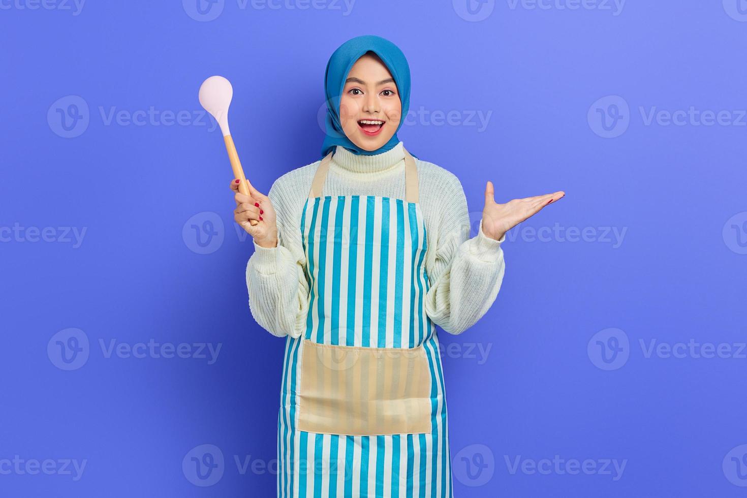 Cheerful beautiful Asian woman in white sweater covered in apron and hijab holding spoon while looking at camera isolated over purple background. Housekeeping concept photo
