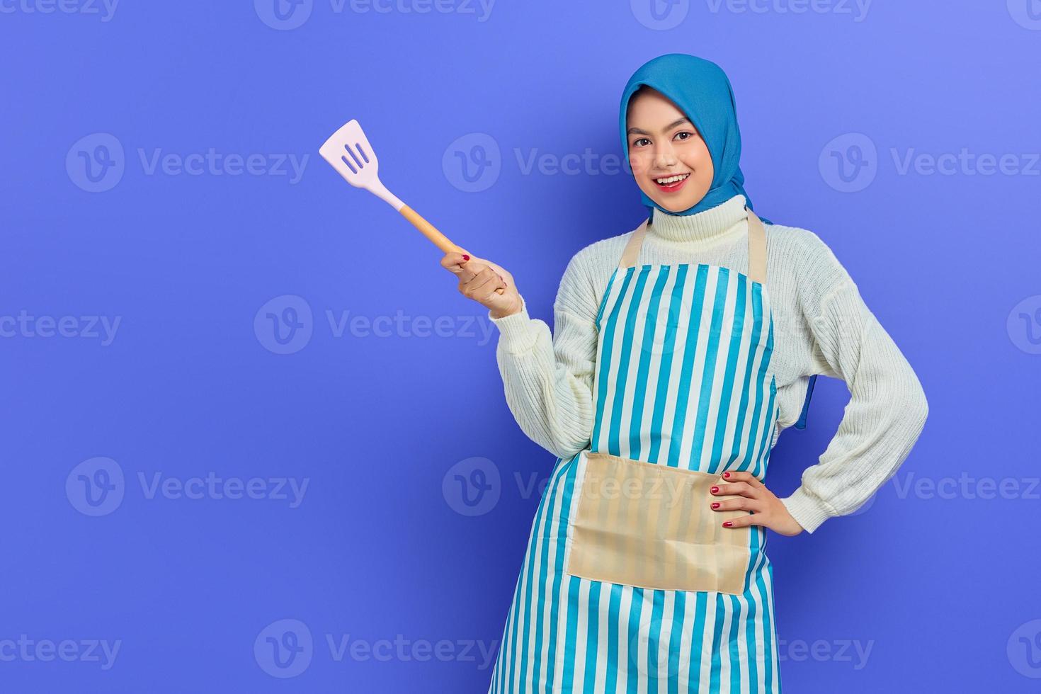 Smiling beautiful Asian woman wearing apron and hijab pointing to copy space with spatula isolated over purple background. Housekeeping concept photo
