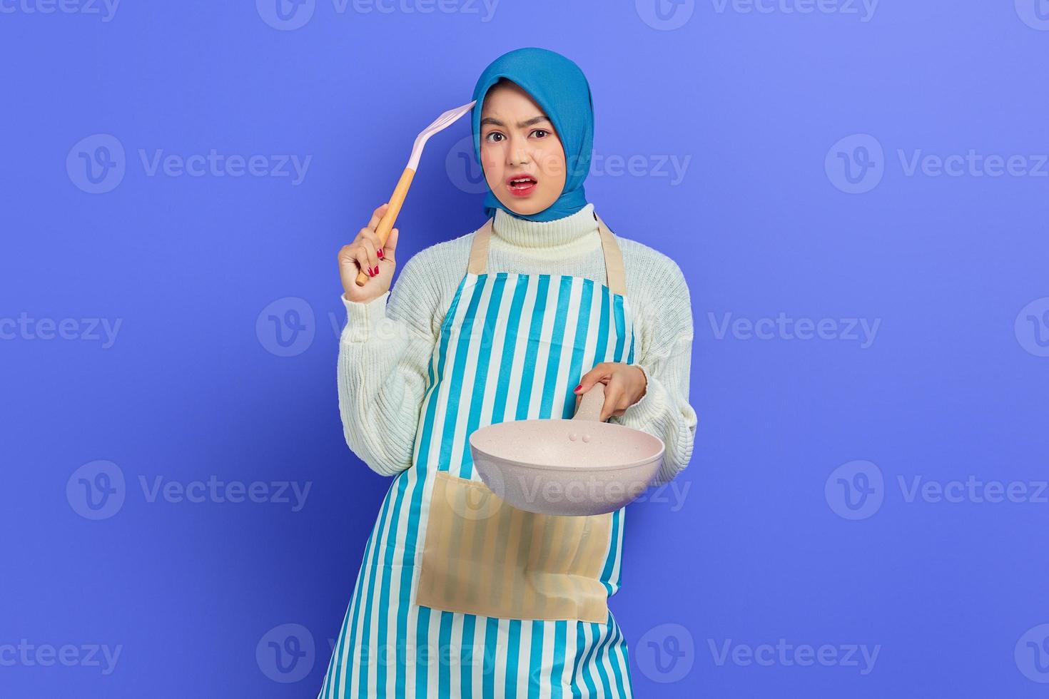 retrato de una joven ama de casa asiática confundida que usa delantal sosteniendo una sartén y una espátula, mirando a la cámara pensando en el menú de cocina aislado sobre fondo morado. concepto de limpieza foto