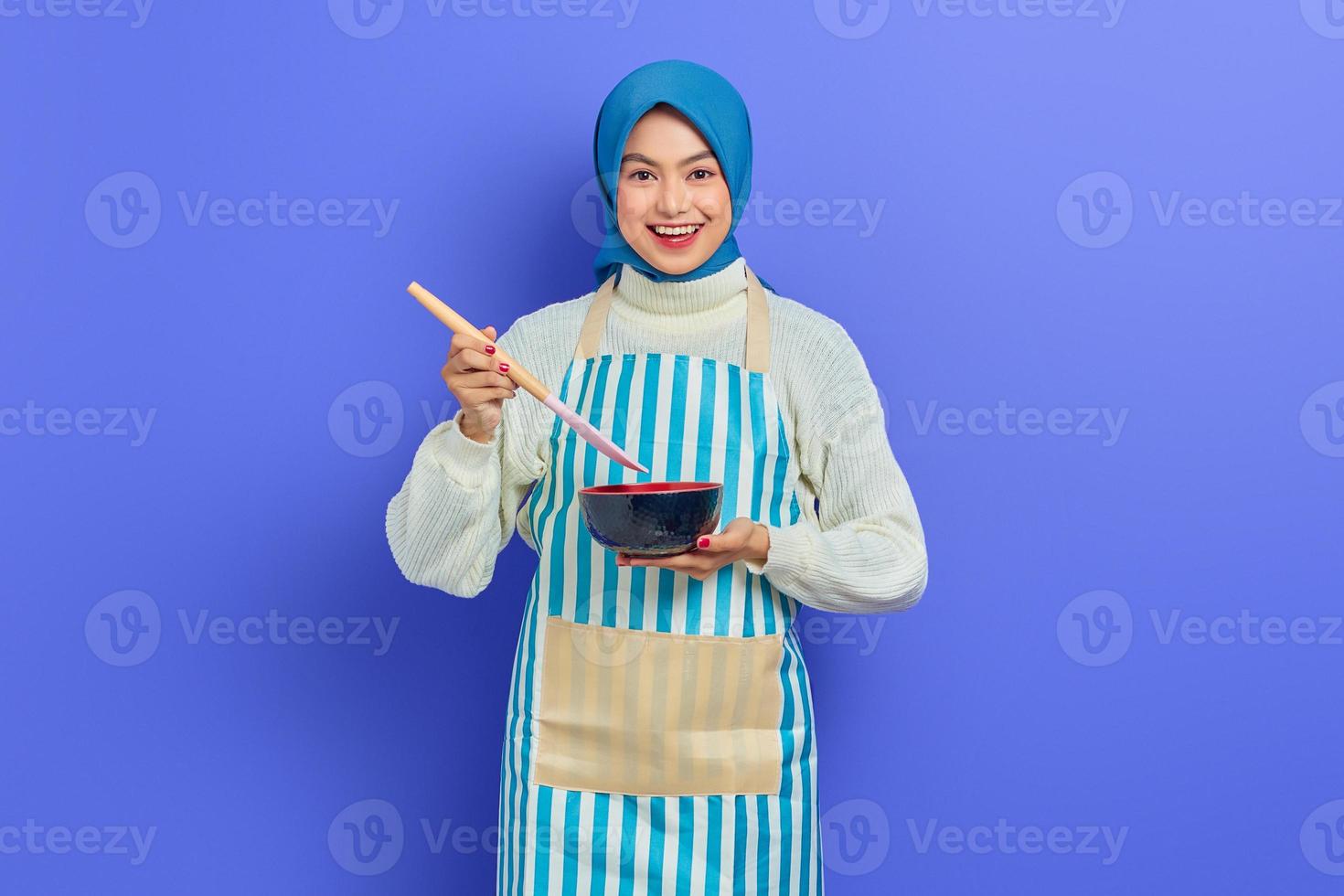 alegre y hermosa mujer asiática con suéter blanco cubierto de delantal y hiyab sosteniendo una cuchara y un tazón mientras hacía las tareas domésticas aisladas sobre un fondo morado. concepto de limpieza foto