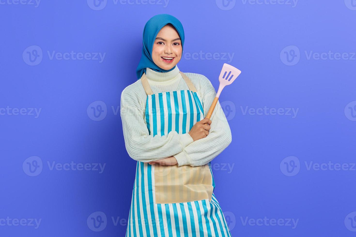 Smiling beautiful Asian woman wearing apron and headscarf holding spatula with crossed arms isolated over purple background. Housekeeping concept photo