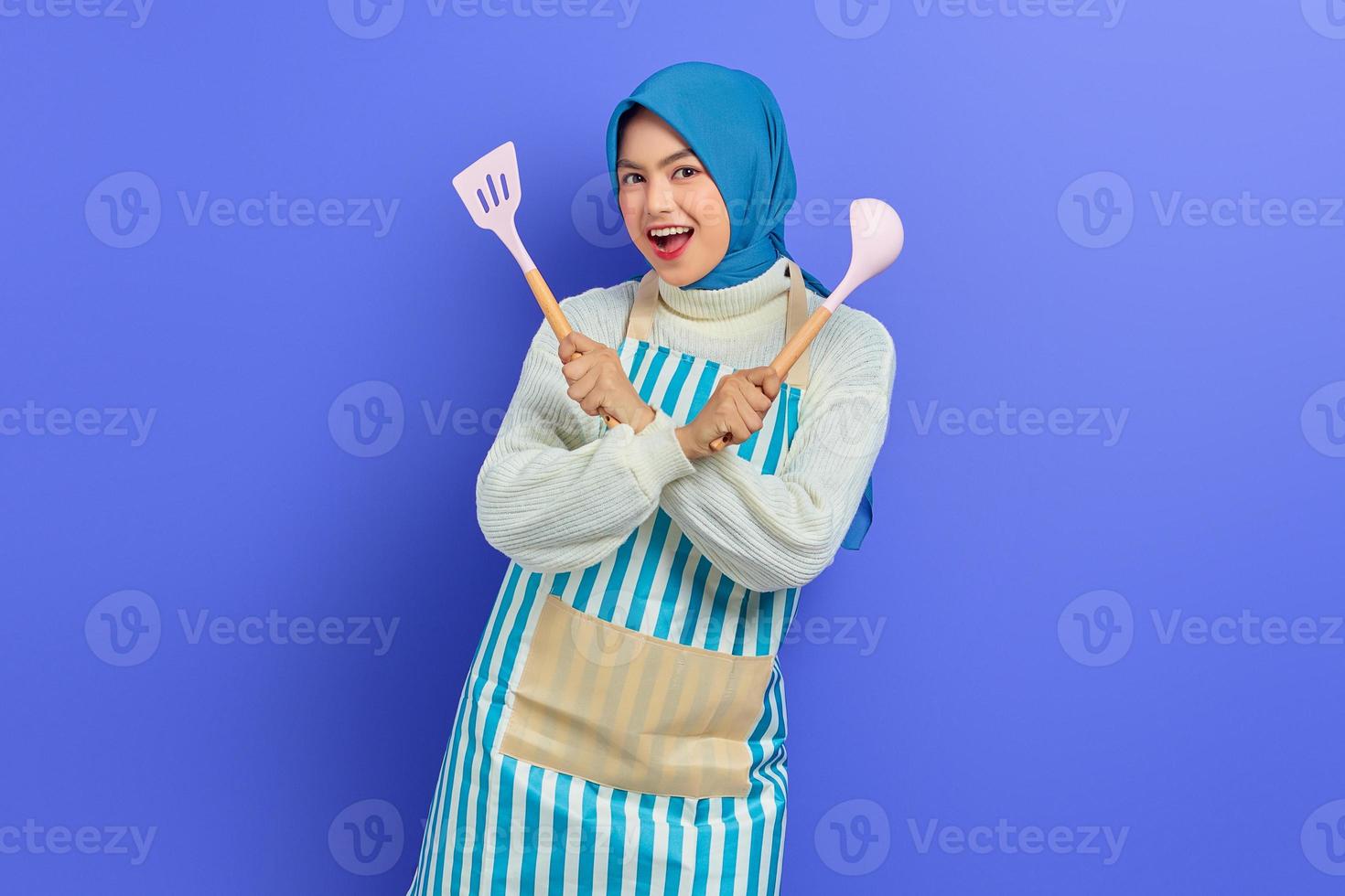 Cheerful beautiful Asian woman in white sweater covered in apron and hijab crossed hand hold spatula and spoon while doing housework isolated over purple background. Housekeeping concept photo