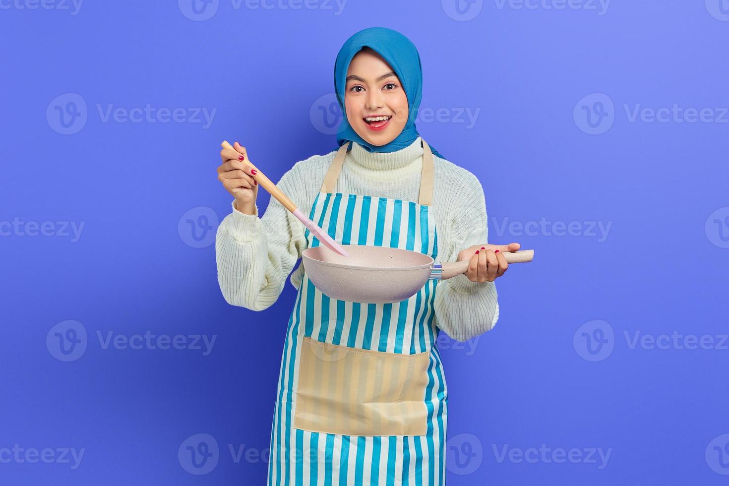 Smiling beautiful Asian woman in white sweater covered in apron and hijab holding spoon and pan while doing homework isolated over purple background. Housekeeping concept photo