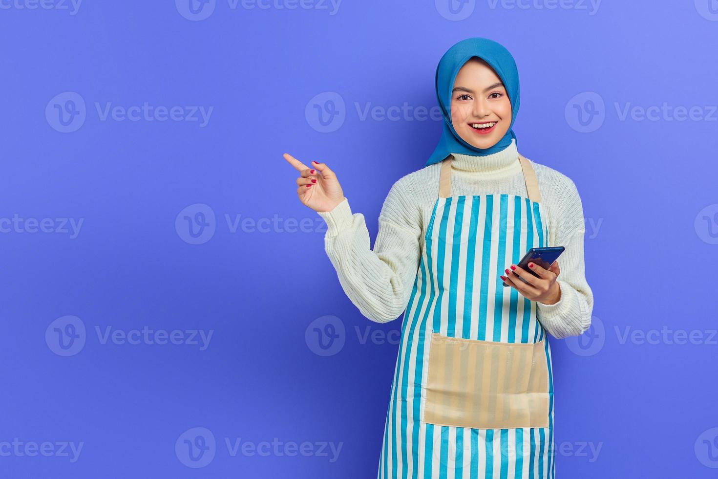 retrato de una joven musulmana asiática sonriente que usa hiyab y delantal, sostiene un teléfono móvil y señala a un lado con los dedos aislados sobre un fondo morado. gente ama de casa concepto de estilo de vida musulmán foto