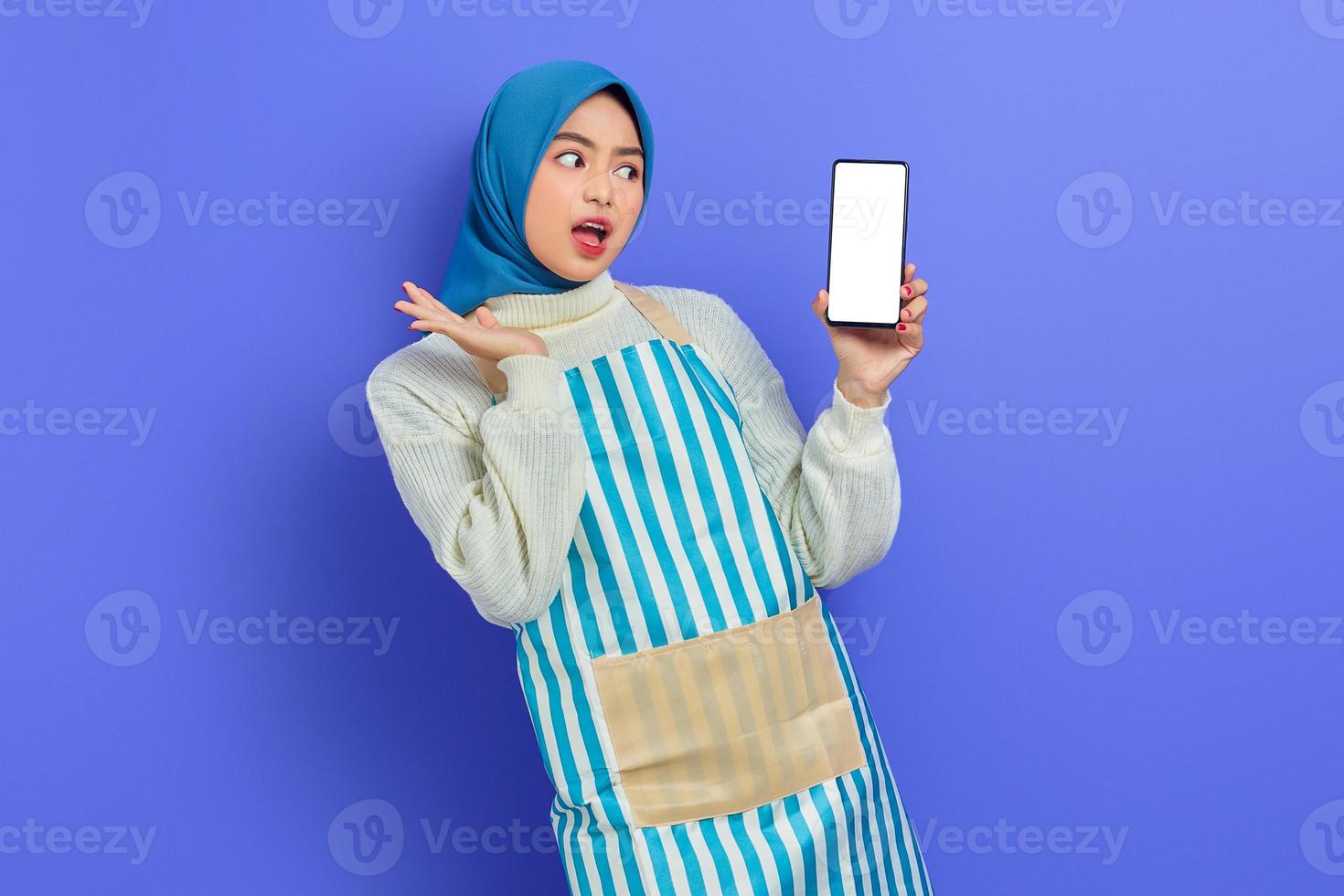 sorprendida joven musulmana asiática usando hijab y delantal sosteniendo un teléfono móvil de pantalla en blanco con la mano aislada en un fondo morado. gente ama de casa concepto de estilo de vida musulmán foto