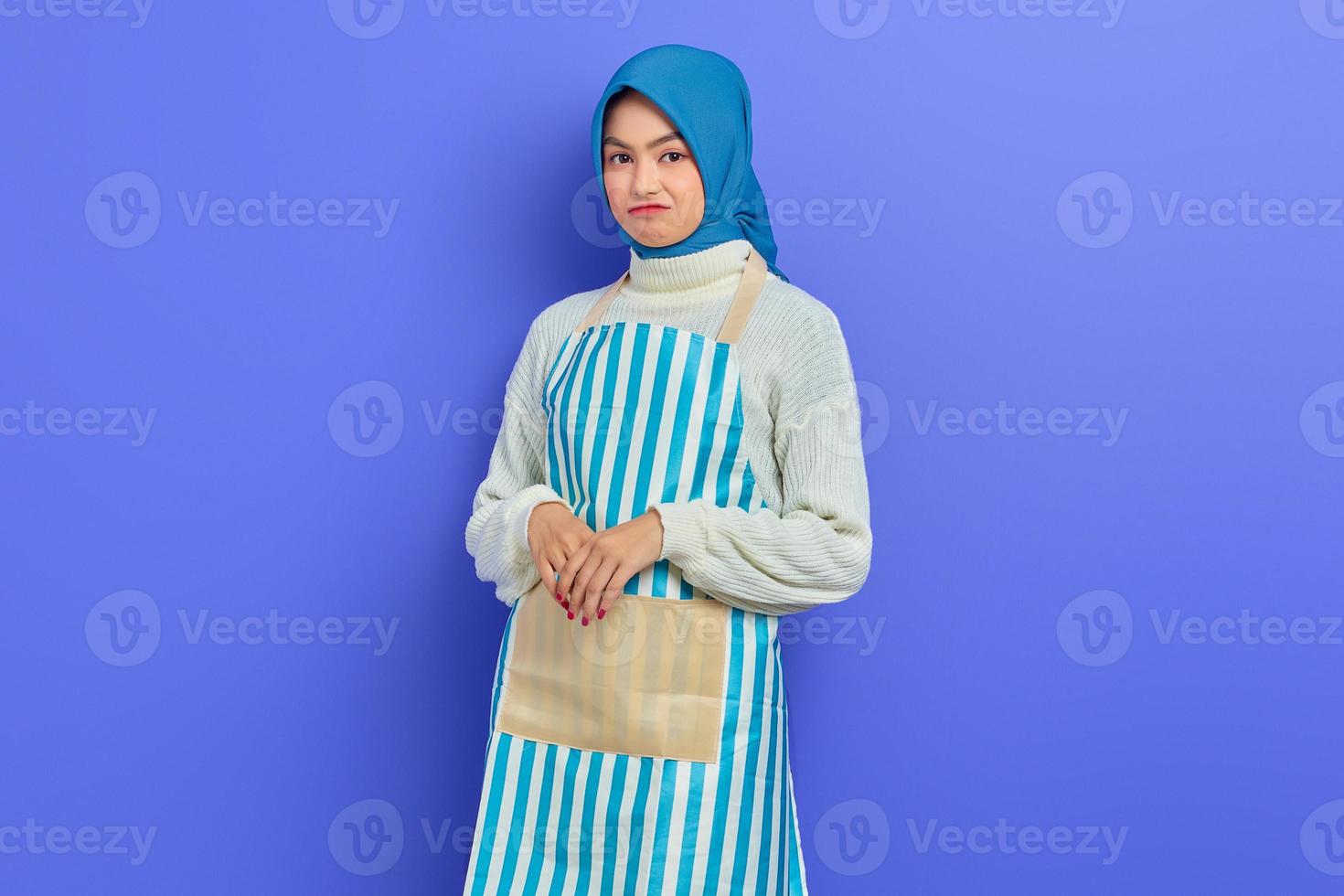 Pensive young Asian Muslim woman in 20s wearing hijab and apron while looking at camera isolated on purple background. People housewife muslim lifestyle concept photo