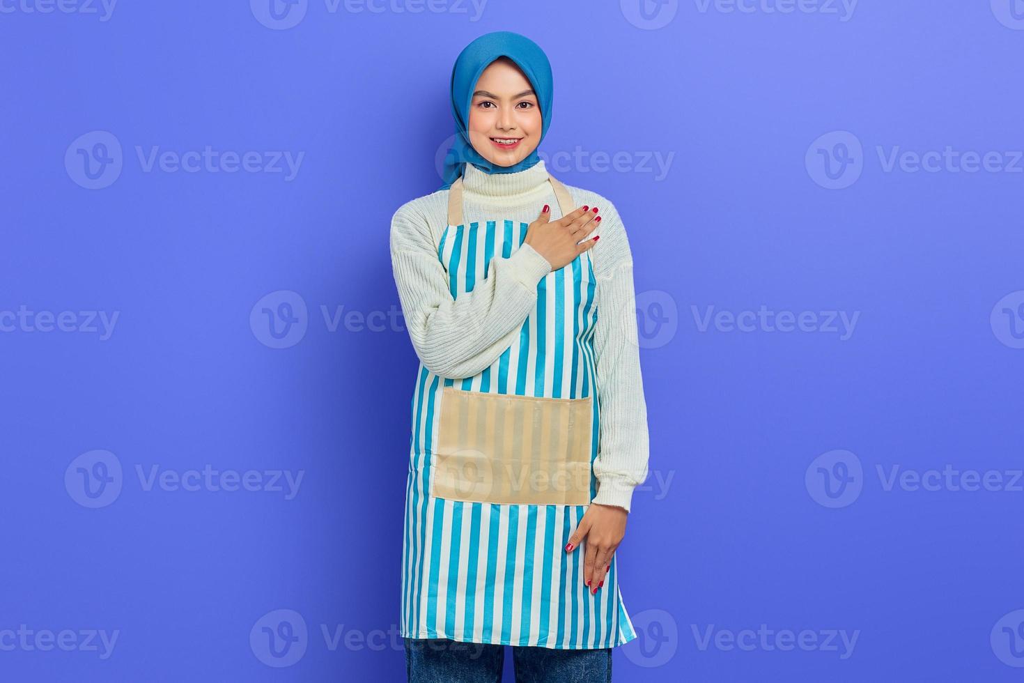 Smiling young Asian Muslim woman in 20s wearing hijab and apron, open sign to greet and greet customers isolated on purple background. People housewife muslim lifestyle concept photo