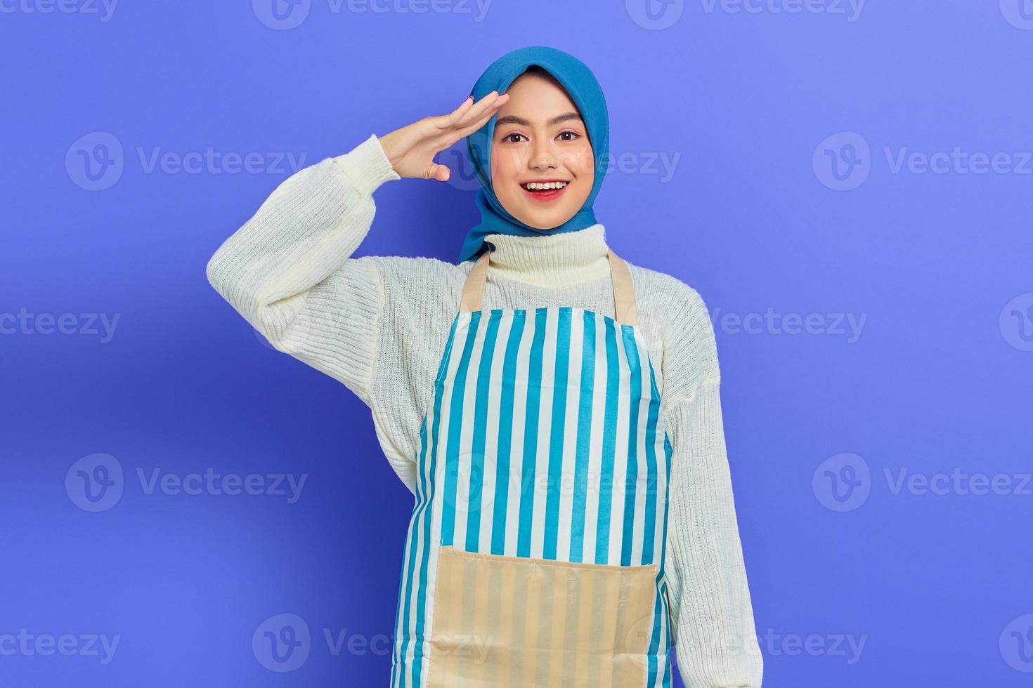 Portrait of excited young Asian Muslim woman wearing hijab and striped apron, showing ready attitude with hands on head while doing homework isolated on purple background photo