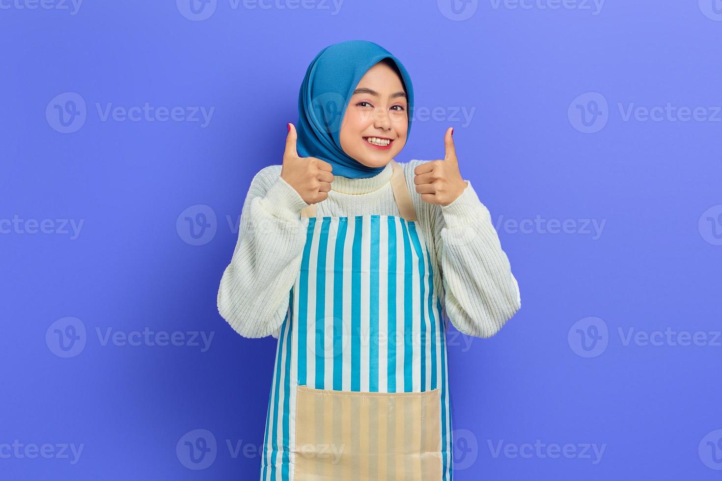 Portrait of cheerful young Asian Muslim woman in hijab and striped apron, standing showing thumbs up and looking camera isolated on purple background. People housewife muslim lifestyle concept photo