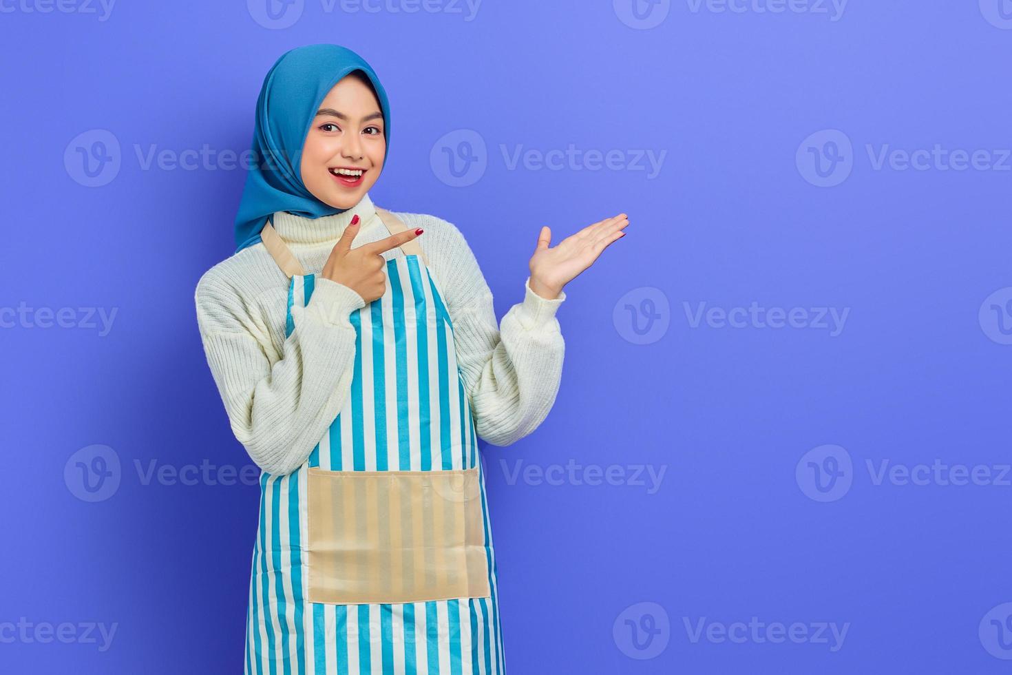 retrato de una joven musulmana asiática sonriente con hiyab y delantal a rayas, mostrando el producto con el dedo y las manos aisladas en un fondo morado. gente ama de casa concepto de estilo de vida musulmán foto