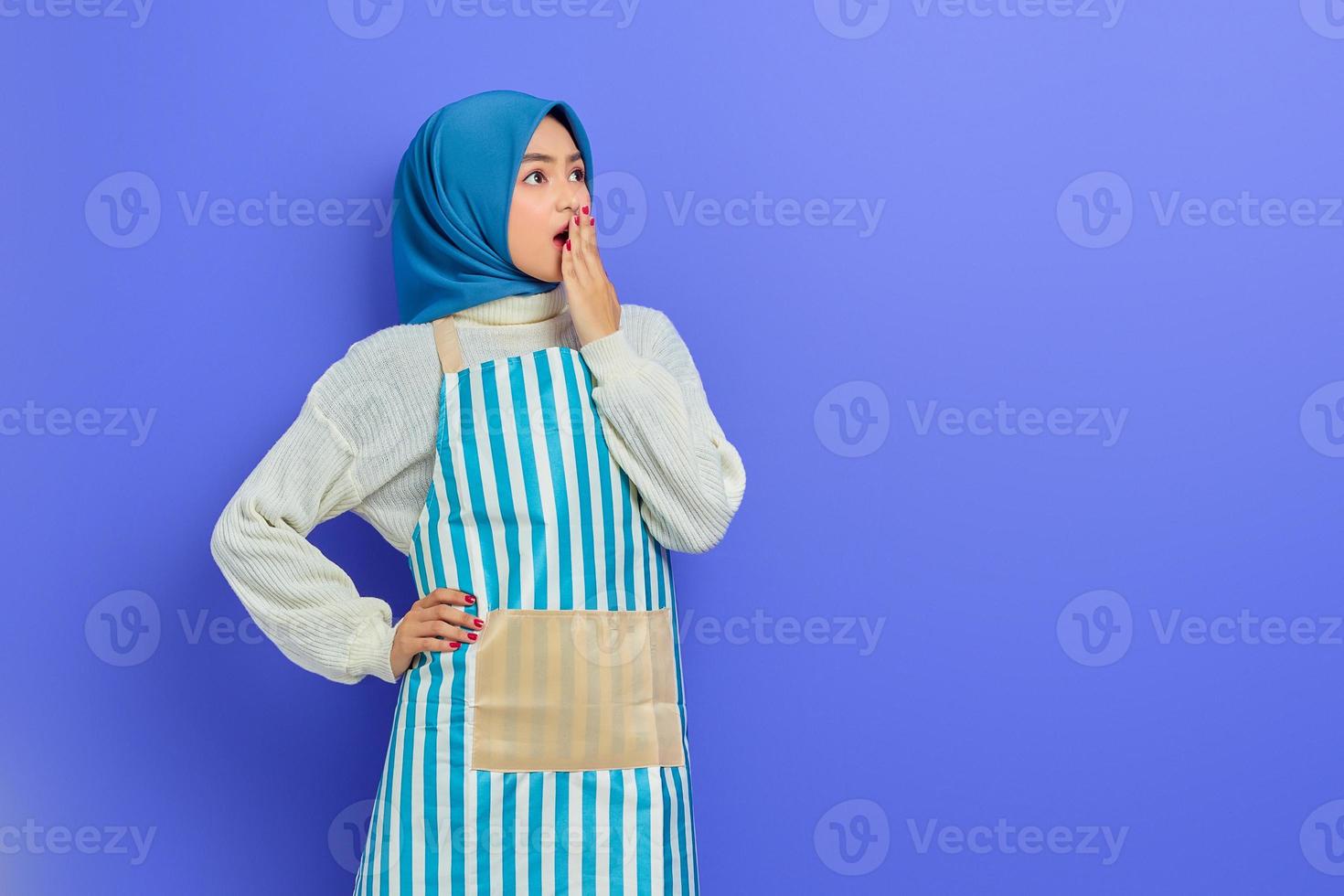 Portrait of shocked young Asian Muslim woman in hijab and striped apron covering mouth with hand, looking aside isolated on purple background. People housewife muslim lifestyle concept photo