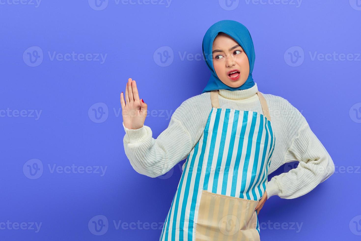 Portrait of angry young Asian Muslim woman in hijab and striped apron showing stop gesture with palm refusing say no isolated on purple background. People housewife muslim lifestyle concept photo